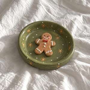May include: A round olive green ceramic trinket dish featuring a raised gingerbread person figure in the centre. The dish is decorated with painted gold starburst patterns and small white dots scattered across the interior surface.