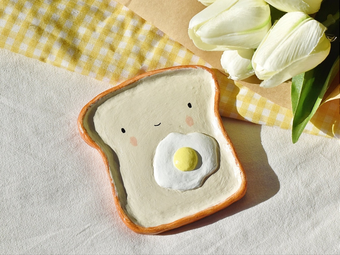 May include: A ceramic dish shaped like a slice of toast with a fried egg on top. The dish is painted with a smiling face and has a yellow yolk and white egg white. The dish is on a white surface with a yellow and white checkered fabric in the background.