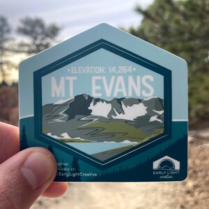 May include: A blue and green hexagonal sticker with a mountain range illustration and the text "ELEVATION: 14,264. MT EVANS" and the logo "EARLY LIGHT creative."