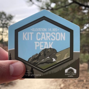 May include: A hexagonal sticker with a blue background and a black border. The sticker features a stylized illustration of Kit Carson Peak, a mountain in Colorado, with the text "• ELEVATION: 14,165. KIT CARSON PEAK" and the logo "EARLY LIGHT CREATIVE".