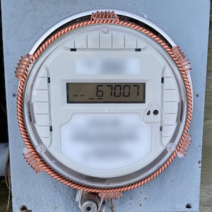 May include: A close-up of a digital electric meter with a copper wire coil around the circular face. The meter displays the number 67007. The meter is mounted on a gray metal box.