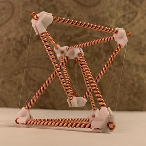 Może przedstawiać: Geometryczna rzeźba z miedzianego skręconego drutu i białych łączników. Struktura to trójwymiarowa forma z wieloma trójkątnymi powierzchniami, połączonymi cienkimi, przezroczystymi sznurkami. Rzeźba jest umieszczona na neutralnym tle.