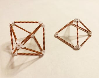 Op de afbeelding: Twee geometrische sculpturen. Eén is een kubusachtige structuur met koperkleurig gedraaid draad en witte connectoren. De andere heeft een ruitvorm met dezelfde materialen. De objecten staan op een wit oppervlak.