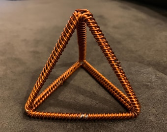 Op de afbeelding: Een handgemaakte piramidesculptuur van koperdraad. De piramide is gemaakt van strak opgerold draad, waardoor een textuur ontstaat. De sculptuur heeft een warme koperkleur en staat op een platte basis. Het is een geometrisch kunstwerk.