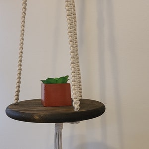 May include: A hanging macrame shelf with a dark brown, round wooden base. A small, square, terracotta-colored pot with a green plant sits on the shelf. The macrame is off-white and hangs from a black hook.