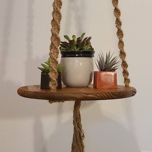 May include: A wooden hanging shelf with three potted plants. The shelf is suspended by a thick rope with a tassel at the bottom. The plants are in different colored pots, including a white pot, a black pot, and a red pot.