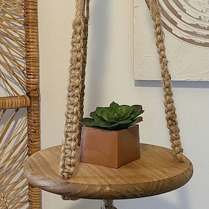 May include: A round wooden hanging shelf suspended by thick braided jute rope. A small square terracotta-colored planter containing a green succulent sits on the center of the shelf. A decorative jute tassel hangs from the bottom of the wooden base.