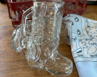 May include: Four clear glass boot-shaped mugs with a decorative design. The mugs are in a red metal rack. The mugs are on a wooden surface.