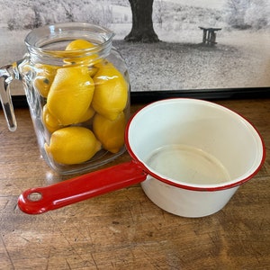 May include: A white enamel saucepan with a red handle. The saucepan is empty and sitting on a wooden surface. A glass jar filled with lemons is in the background.