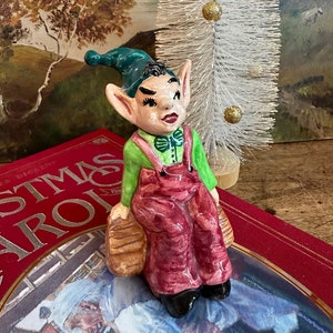 May include: A ceramic elf figurine with green pointed ears, a green hat, and a green shirt. The elf is wearing red overalls and is sitting on a brown barrel. The elf has a painted expression with black eyes, black eyebrows, and a red mouth.