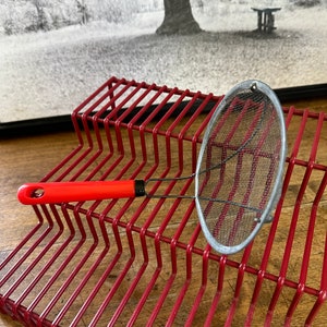 May include: A red metal rack with a small, silver, mesh strainer with a red handle.