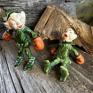 May include: Two vintage ceramic elf figurines. The elves are dressed in green outfits with orange gloves. One elf is standing with closed eyes, and the other is lying down with an open mouth. The figurines are on a weathered wooden surface.