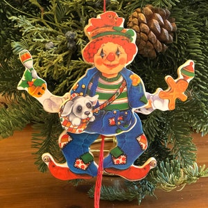 May include: A wooden pull-string toy ornament of a clown with a red hat, blue pants, and a white dog. The clown is holding a gingerbread cookie and a red bird. The ornament is hanging from a red string.