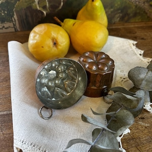May include: A still life arrangement featuring ripe yellow pears, two antique metal molds, and sprigs of eucalyptus on a linen cloth. The molds are detailed with floral patterns. The scene is set on a wooden surface.