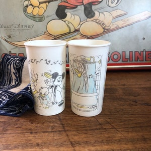 May include: Two white paper cups with cartoon images of Mickey Mouse and Donald Duck. The cups are on a wooden surface. The image in the background is of Mickey Mouse skiing.