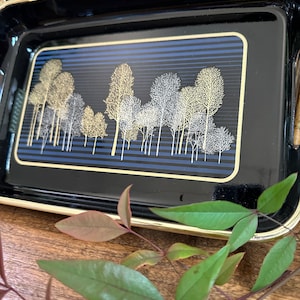 May include: A black serving tray with a gold rim and a blue and gold design of trees. The tray is sitting on a wooden surface.