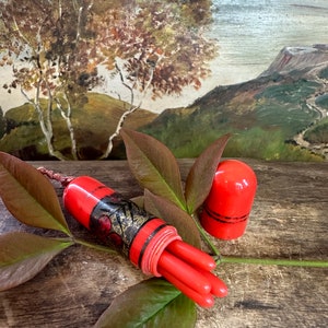 May include: A red and gold vintage sewing thimble with a floral design. The thimble is open and five red sewing needles are inside. The thimble is sitting on a wooden surface with green leaves.