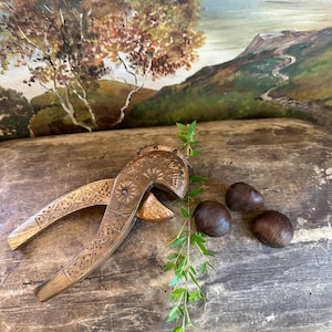 May include: A wooden nutcracker with intricate carvings, resting on a rustic wooden surface with three brown chestnuts. A sprig of green leaves adds a touch of nature to the scene.