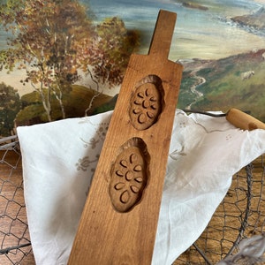 May include: A wooden cookie mold with two carved floral designs. The mold is brown and has a handle. It is sitting on a white cloth and a wire basket.