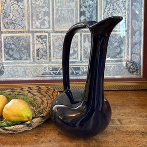 May include: A dark blue ceramic vase with a unique, curved design. The vase has a wide base and a narrow neck, with a spout at the top.
