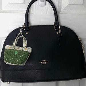 May include: Black leather handbag with a top handle and a small, decorative fabric bag attached. The small bag has a green polka dot pattern and a white daisy print. The handbag has a gold Coach logo.
