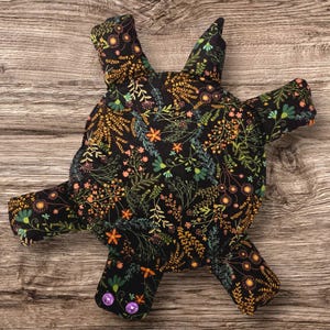 May include: A stuffed fabric toy shaped like a turtle with a black background featuring a colorful floral pattern of orange, yellow, and green plants. The toy has four legs, a head, and a tail, with two small purple circular snaps on one leg.