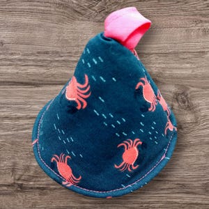 May include: A teal fabric bib with a pink trim and a pink loop at the top. The bib is patterned with coral-colored crab illustrations and white dashes. The bib is designed to catch spills and protect clothing.