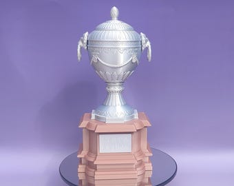 May include: A silver-colored decorative urn with a lid, featuring ornate detailing and handles. The urn sits atop a pink rectangular base with the text "COPPA LATINA" on a plaque. The background is a solid purple color.