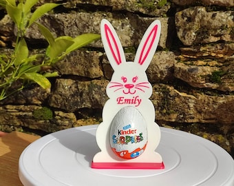 May include: White wooden bunny rabbit Easter egg holder with pink ears and a pink base. The bunny has a pink nose and eyelashes. The bunny is holding a silver foil wrapped Kinder Surprise chocolate egg. The bunny has the name "Emily" written on it.