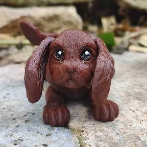 Puede incluir: Una pequeña figurita de cachorro marrón con orejas grandes y grandes ojos marrones. La figurita está hecha de un material texturizado y tiene una apariencia realista.