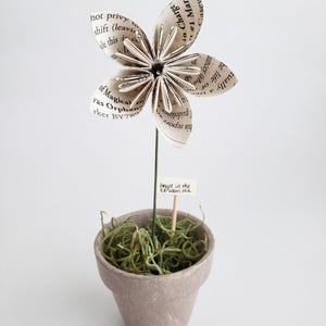 Puede incluir: Una flor de papel hecha de una página de libro, con un tallo verde y un pequeño letrero que dice "House in the cerulean sea", está plantada en una maceta marrón con musgo verde.