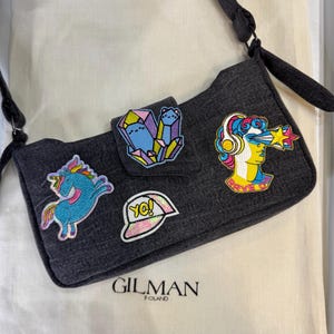 Könnte beinhalten: Dunkelgraue Denim-Umhängetasche mit schwarzem Riemen, verziert mit bunten gestickten Aufnähern. Aufnäher sind ein Einhorn, Kristalle, eine Mütze mit "Yo!" und eine Frau mit Kopfhörern. Die Tasche ist von "GILMAN POLAND".
