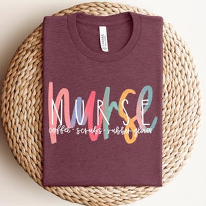May include: A maroon t-shirt with the word "nurse" in colorful, stylized lettering. Below the word "nurse" are the words "coffee, scrubs, rubber gloves" in a smaller font. The shirt is displayed on a woven, circular surface.