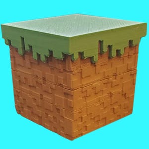 May include: A square, Minecraft-themed box with a green top and a brown, block-like design on the sides. The top edge has a jagged, grass-like appearance. The box is set against a bright blue background.