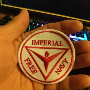 May include: White embroidered patch with a red triangle and a red flame inside. The triangle has the words "IMPERIAL FREE NAVY" written around it.