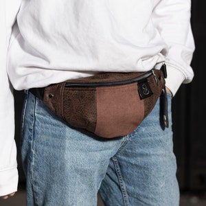 Könnte beinhalten: Eine braune Leder-Bauchtasche mit einem Reißverschlussfach und einem braunen Leder-Patch mit einem Logo. Die Bauchtasche wird um die Taille einer Person getragen, die eine blaue Jeans trägt.