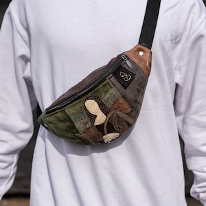 May include: A brown, green and beige patchwork fanny pack with a zipper closure and an embroidered image of the Mona Lisa. The fanny pack has a black adjustable strap.