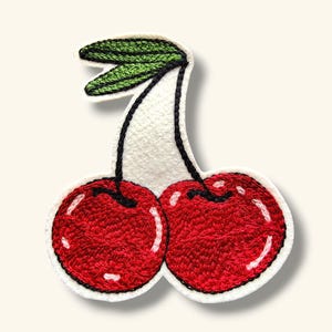 May include: A pair of red cherries with green stems and leaves, embroidered on a white fabric background.