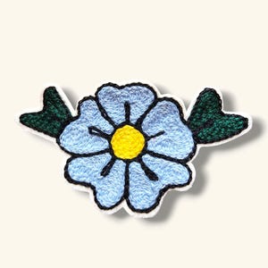May include: A blue embroidered flower with a yellow center and green leaves. The flower is surrounded by a white background.