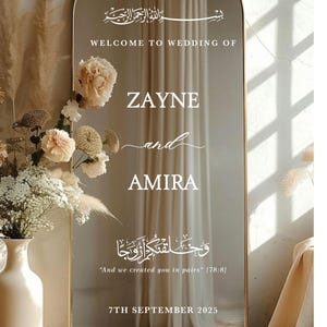 Op de afbeelding: Een grote, boogvormige spiegel met een gouden rand, met witte tekst: "WELCOME TO WEDDING OF ZAYNE and AMIRA" en de datum "7TH SEPTEMBER 2025." De spiegel is versierd met bloemstukken en een vaas.