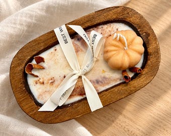 May include: An oval wooden dough bowl filled with white wax, featuring a light orange pumpkin-shaped candle, cinnamon sticks, and a white ribbon tied in a bow with the printed text BEST WISHES FOR YOU.