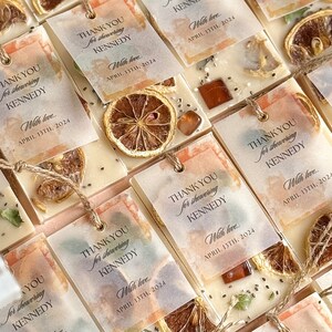Personalized burnt orange soy wax sachet favors featuring dried orange slices, handmade soy wax air fresheners and custom vellum labels with names and dates. Elegant wedding favors, bridal shower favors, baby shower gifts, engagement party favors and wedding guest favors. Neutral boho decor with natural botanicals and premium fragrance oils. Perfect for wedding welcome bags, bridesmaid gifts, bridal events and special celebrations.