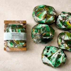 Puede incluir: Jabón de piedra de eucalipto con un diseño de mosaico en tonos verdes, blancos y dorados. Un jabón está en una jabonera de madera con la etiqueta "Eucalyptus stone soap". Otros jabones están dispersos.