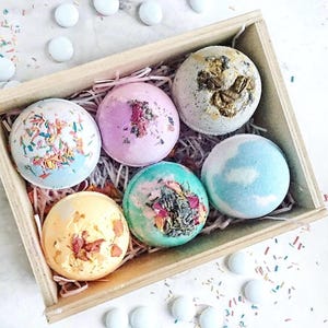 Puede incluir: Una caja de madera llena de seis bombas de baño coloridas. Las bombas de baño son redondas y están decoradas con flores secas y chispas. La caja está sobre una superficie blanca con confeti esparcido y objetos redondos blancos.