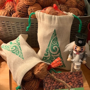 May include: Two burlap bags with green Christmas tree designs filled with walnuts. A nutcracker figurine stands next to the bags. A business card for Nutty Acres Nuts reads "We're nutz about our nuts!" with a website address.