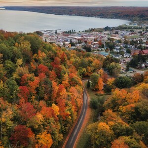 May include: Aerial view of a small town nestled in a valley surrounded by a vibrant forest of red, orange, and yellow fall foliage. A winding road leads up to the town, which is situated on the edge of a large lake.