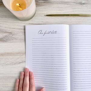 May include: An open journal with lined pages and the text "Our first date..." written on the left page. A hand is holding the journal open. A gold pen and a white candle are on a wooden surface.