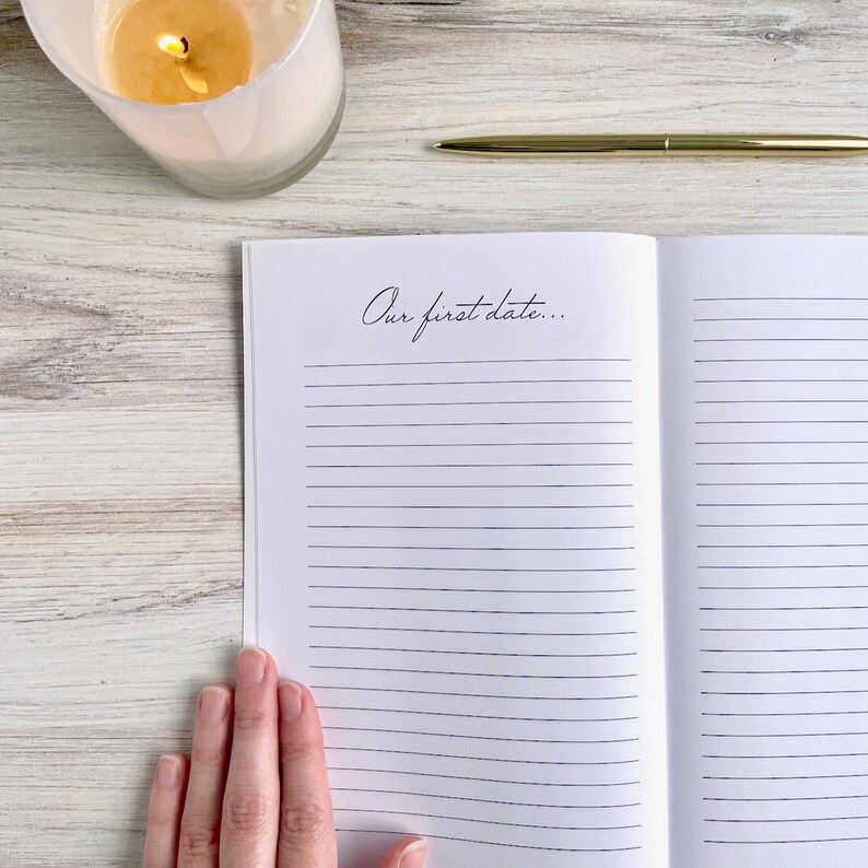 May include: An open journal with lined pages and the text "Our first date..." written on the left page. A hand is holding the journal open. A gold pen and a white candle are on a wooden surface.