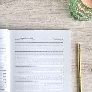 May include: A white lined notebook with the text "Days Until Wedding" and "Date" written on the top of the page. A gold pen is lying on a white wooden surface next to the notebook.