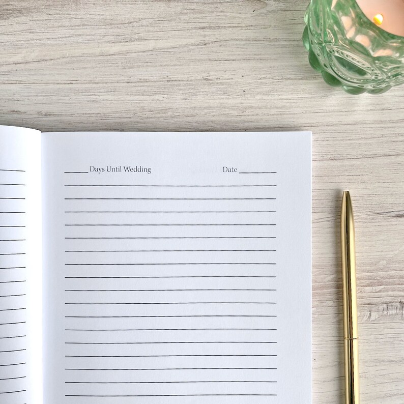 May include: A white lined notebook with the text "Days Until Wedding" and "Date" written on the top of the page. A gold pen is lying on a white wooden surface next to the notebook.